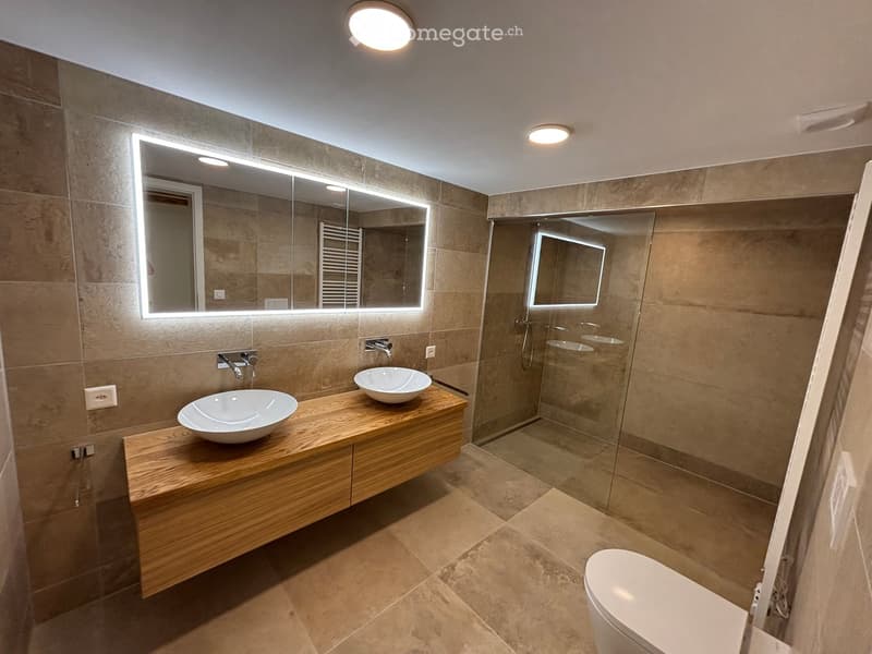 Modern bathroom with wooden vanity, two sinks, large illuminated mirror, and glass-enclosed shower area.
