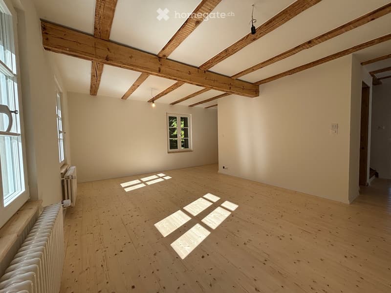 Empty room with wooden floor, exposed wooden beams, white walls, and natural light from windows.