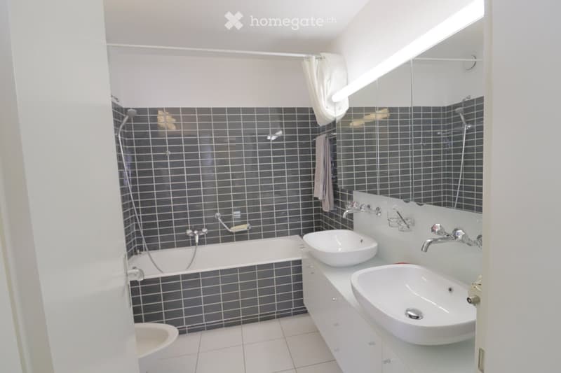 Modern bathroom with dark tiled walls, white bathtub, double sink vanity, large mirror, and towel hanging on the wall.