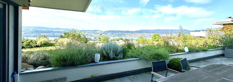 Balcony with outdoor seating, overlooking a scenic view of a lake and distant mountains, surrounded by greenery and plants.