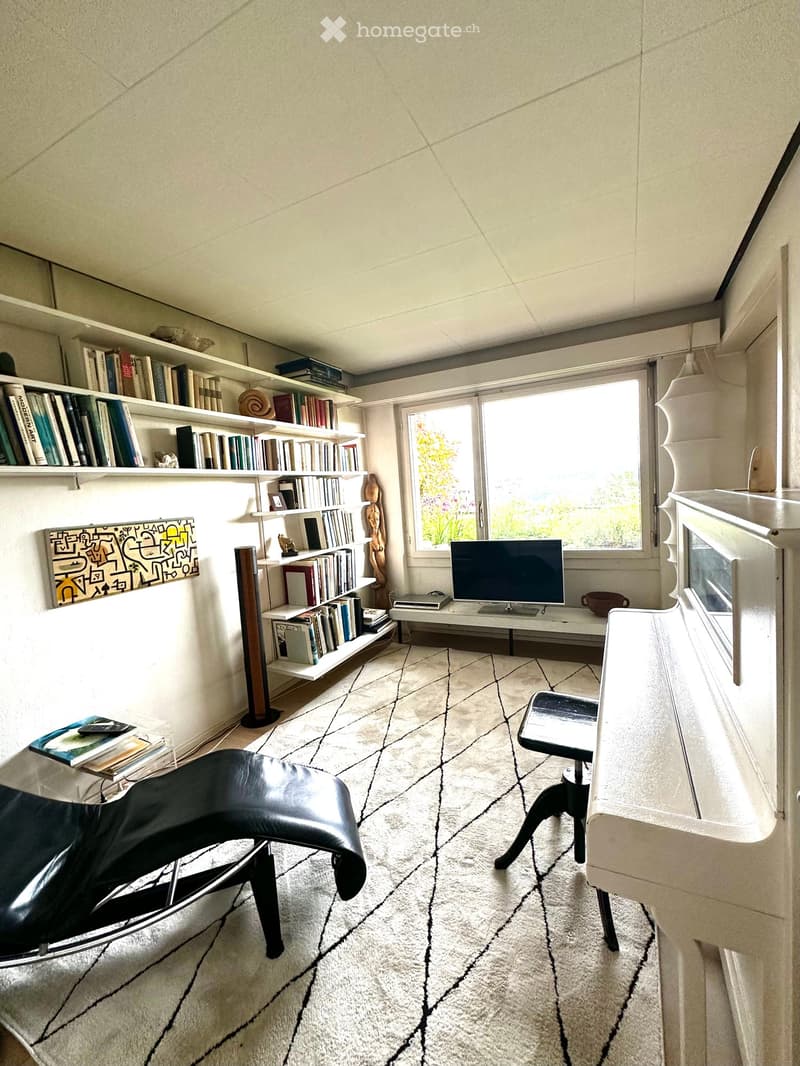 Modern, well-lit room with a large window, featuring a black leather chair, a white table with a screen, a bookshelf filled with books, a piano, and a patterned rug.