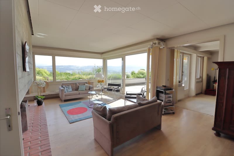 Bright living room with large windows offering scenic views, featuring a sofa, chairs, a coffee table, a lamp, a colorful rug, and a flower vase.