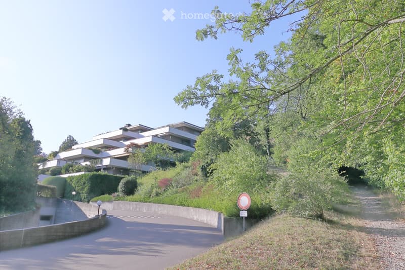 Modern multi-story residential building with white facade, surrounded by lush greenery and trees. The building features balconies and is situated on a raised ground floor with a curved driveway leading up to it.