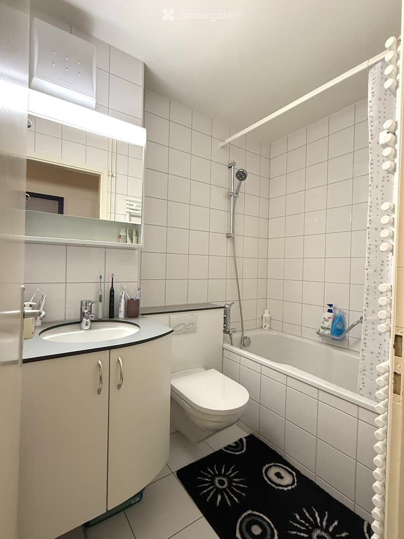 Modern bathroom with white tiles, a sink with a mirror cabinet, a toilet, a bathtub with a shower, and a black bath mat with a sun design.