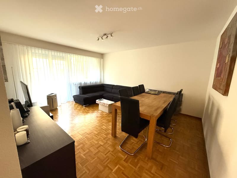 A modern living room with a dark wooden floor, a black sectional sofa, a wooden dining table with chairs, a TV screen, and a small table with a stool.