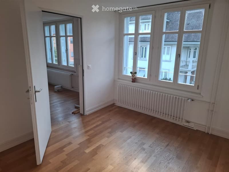 Empty room with wooden flooring, large windows, radiators, and a small potted plant on the windowsill.