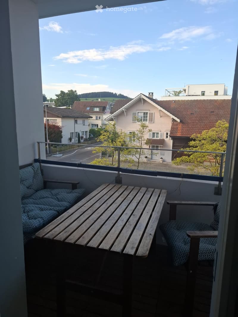 Balcony with wooden table and two chairs, overlooking a residential area with houses and trees.