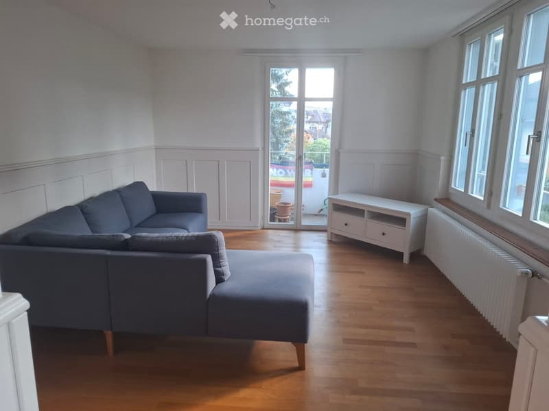 Bright living room with wooden flooring, large windows, a grey sectional sofa, ottoman, and a white TV stand.