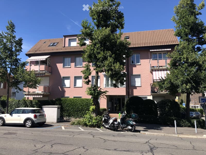 Two-story house with pink brick facade, multiple windows, balconies, and a tiled roof. The house is surrounded by greenery and has a small entrance area with steps.
