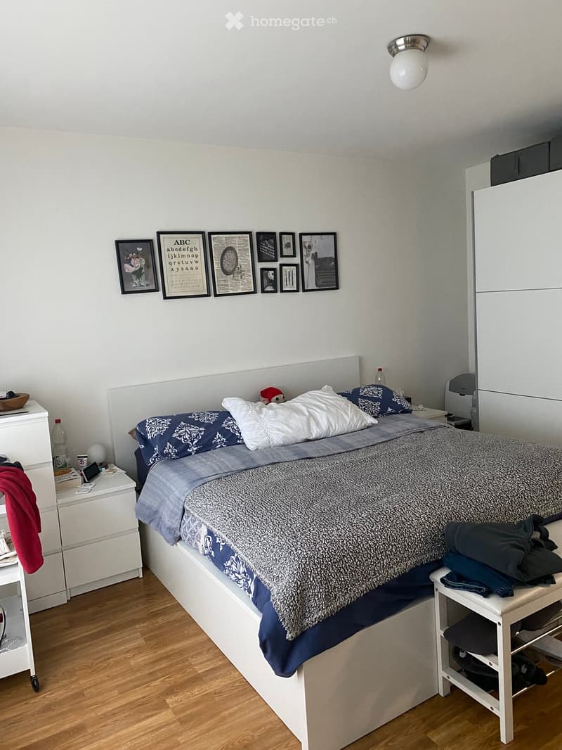 A modern bedroom with a large bed, nightstand, wardrobe, and framed pictures on the wall. The bed has a blue and white patterned duvet and pillows. A small stool holds folded clothes. A mirror is mounted on the wardrobe.