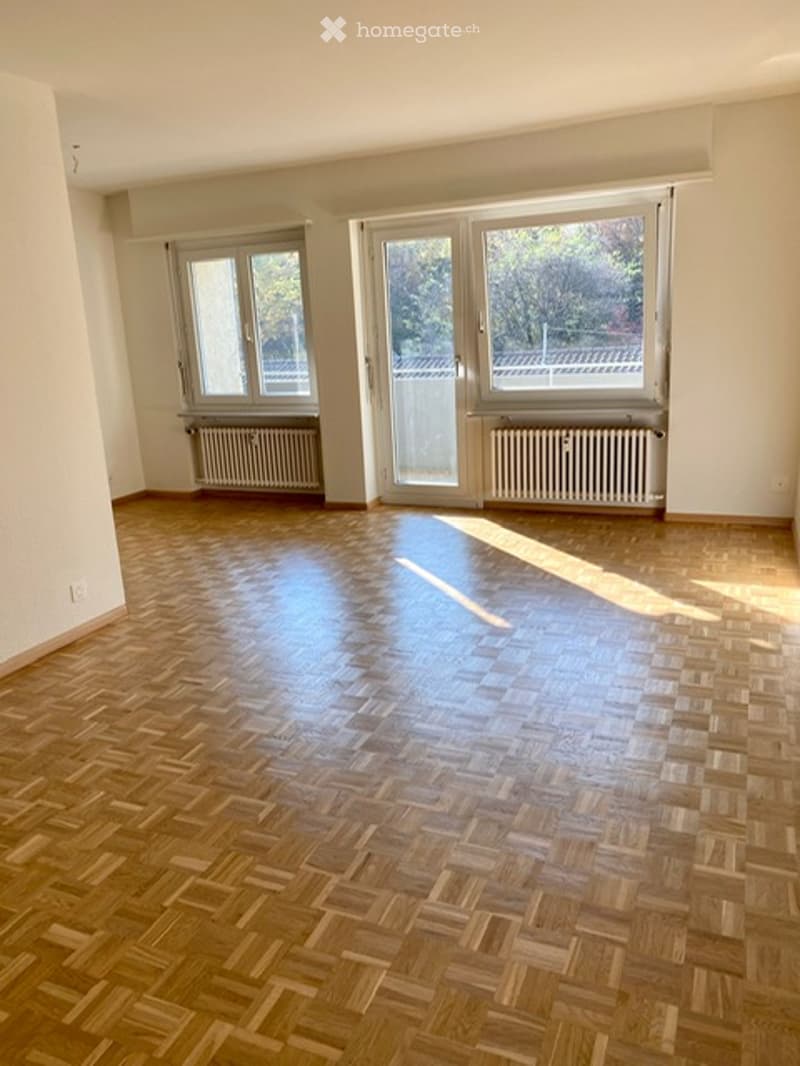 Empty room with wooden parquet flooring, large windows, radiators, and natural light.
