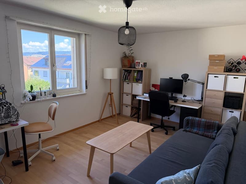 A modern, well-lit living and working space with a sofa, coffee table, desk, laptop, bookshelf, and storage unit. The room features large windows with a city view and is furnished with a mix of contemporary and functional pieces.