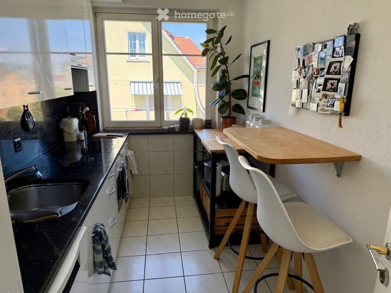 Modern kitchen with white cabinets, black countertops, and a small dining area with stools and a table. Features a large window with a view of the building outside.