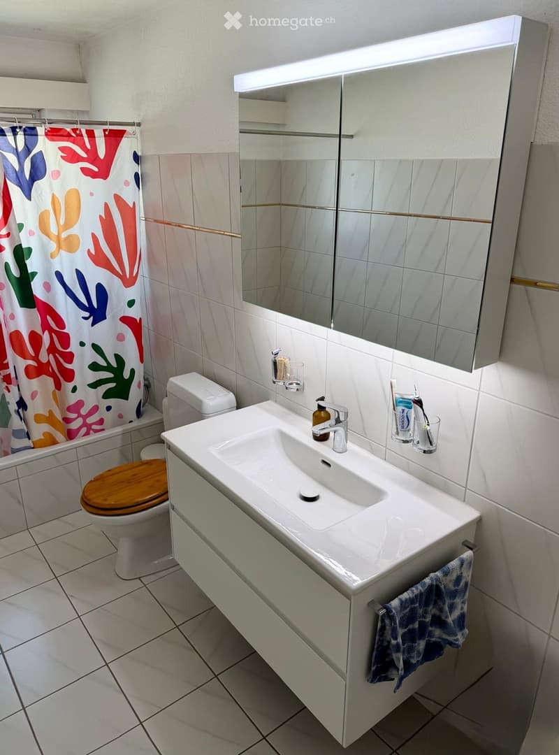 Modern bathroom with white tiles, a white sink with a modern faucet, a large mirror with built-in storage, a toilet with a wooden seat, and a colorful shower curtain.