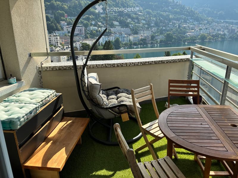 Balcony with a scenic view of a lake and town, featuring a wicker hanging chair, wooden table, wooden chairs, and a bench.