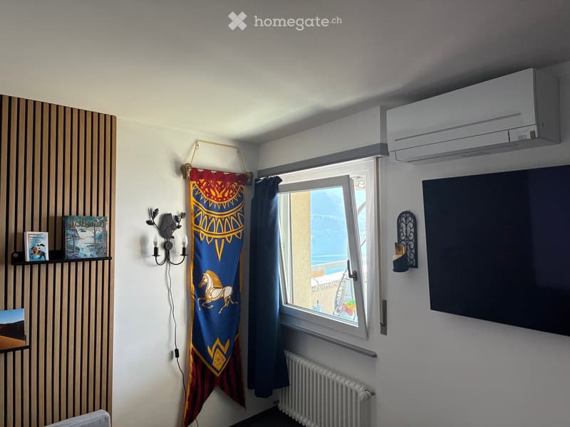 A furnished room with a large window offering a view, a shelf with books, a wall-mounted light fixture, a decorative curtain, a radiator, an air conditioner, and a screen on the wall.