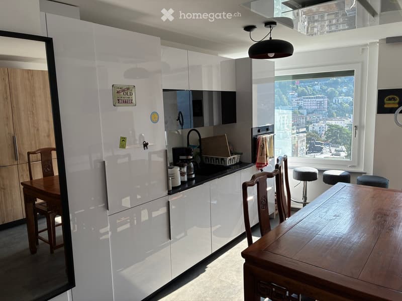 Modern kitchen with white cabinets, a large window offering a city view, a wooden dining table, and stools. Includes a refrigerator, coffee maker, and various kitchen items on the counter.