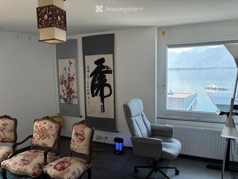 A modern living room with a view of a lake and mountains, featuring a sofa, several chairs, a table lamp, and art pieces on the wall.