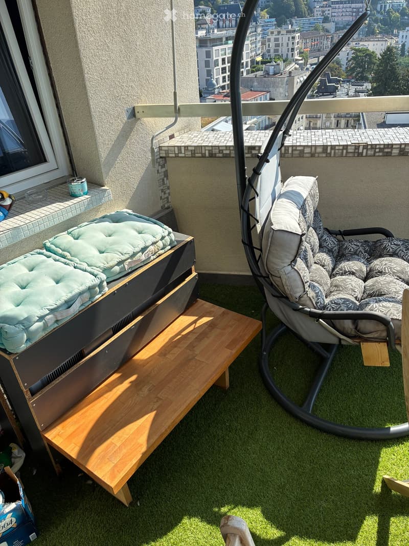Balcony with artificial grass, swing chair, wooden bench, and pet bed, overlooking a cityscape.