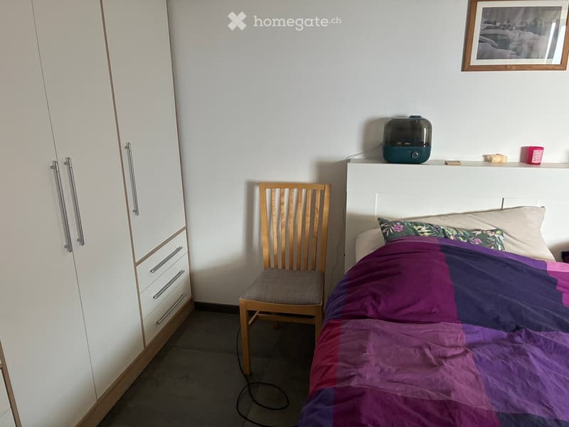 A tidy bedroom with a bed covered in a purple and pink duvet, a wooden chair, a white wardrobe, and a picture frame on the wall.