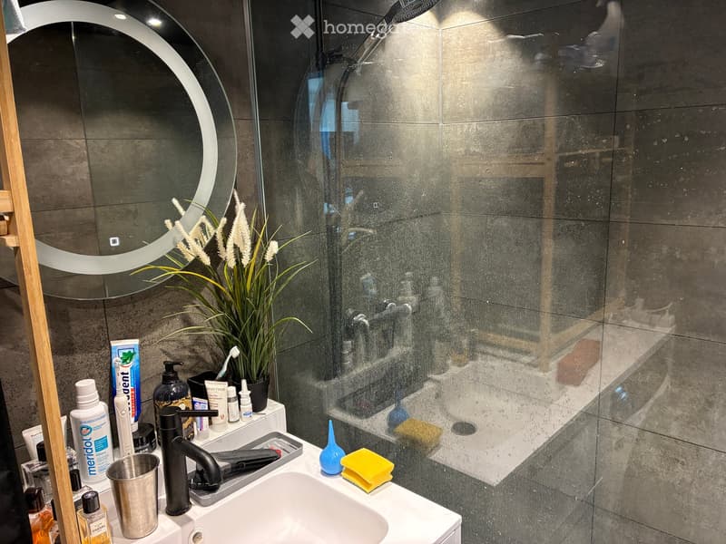 Modern bathroom with a white sink, black faucet, round mirror, and a glass-enclosed shower. The countertop holds various toiletries, a plant, and a yellow sponge.