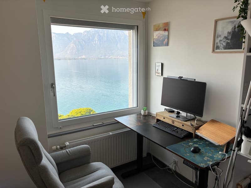 A modern home office with a large window overlooking a lake and mountains, featuring a desk with a monitor, keyboard, mouse, and a comfortable chair.