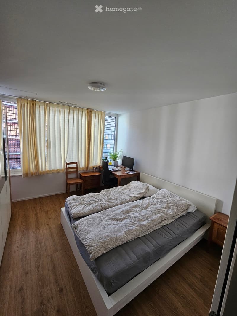 A tidy bedroom with a double bed, a wooden desk with a laptop, a chair, and a small plant near the window. The room features light-colored curtains and a wooden floor.
