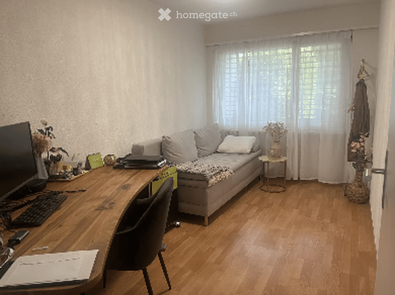 A small, well-lit living room with a grey sofa, a wooden desk with a laptop, a chair, and a small side table with a vase. The room has wooden flooring and large windows with white curtains.