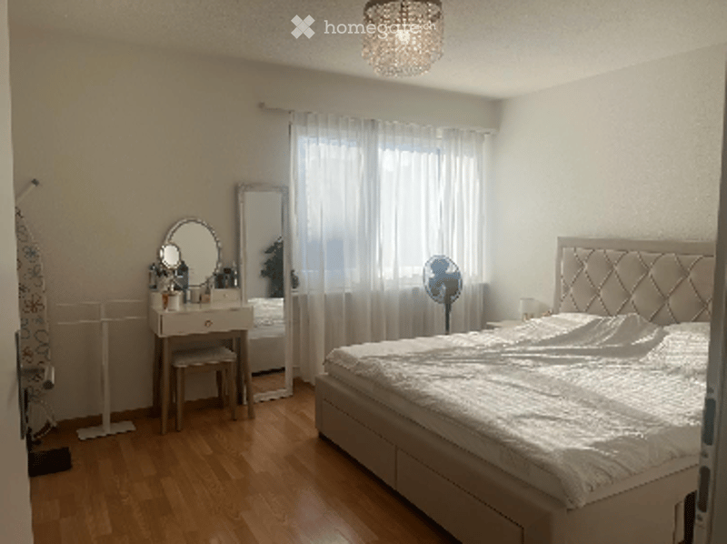 Bright bedroom with wooden flooring, large window with white curtains, a tufted headboard bed, a vanity with a mirror, and a small fan near the window.