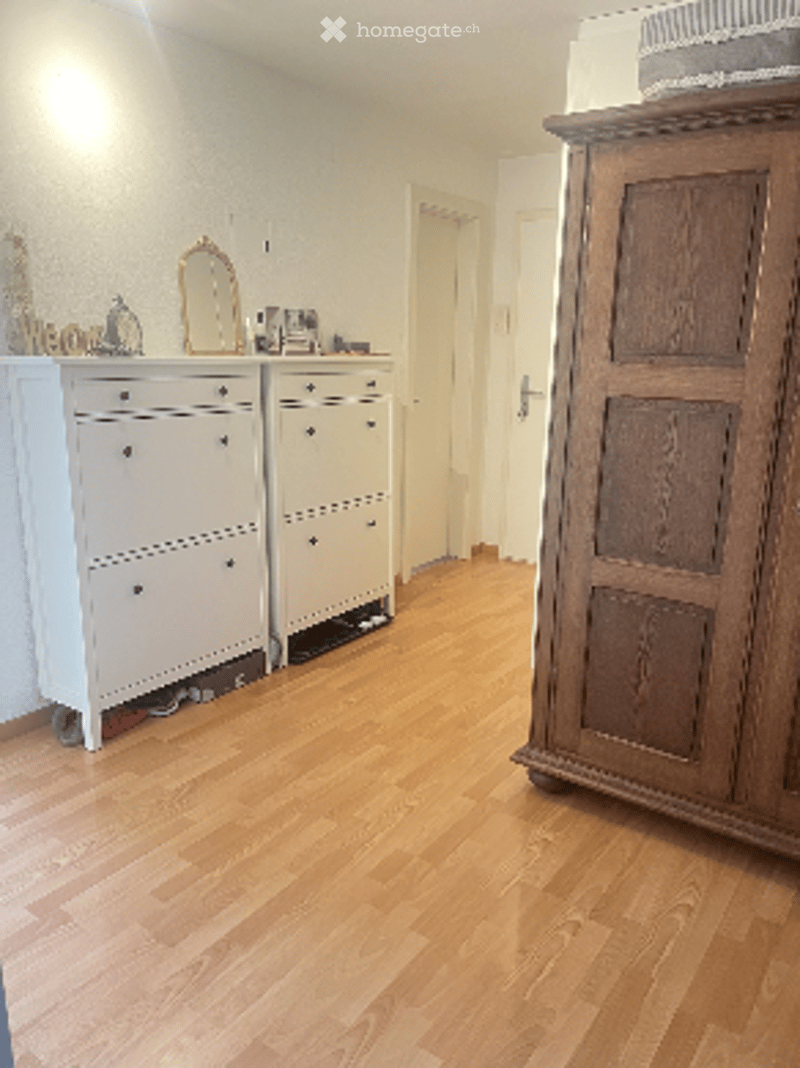 A well-lit room with light wooden flooring, a large wooden wardrobe, a white drawer unit, a mirror, and a small picture frame on top of the drawer unit.