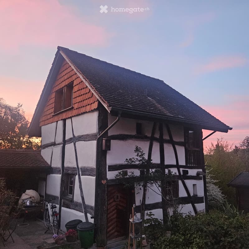 Traditional half-timbered house with white plaster walls and dark wooden beams, featuring a steeply pitched roof with red tiles and multiple windows. The house is surrounded by greenery and has a small garden area with potted plants and a bicycle near the