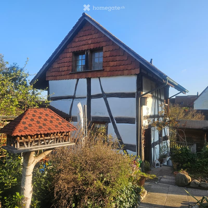 Traditional half-timbered house with white plaster walls and red-tiled roof, surrounded by garden and small wooden structure.