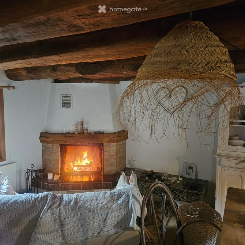 Cozy living room with exposed wooden beams, a lit fireplace, a sofa with striped cushions, a small table with candles and decorative items, and a woven basket on the floor.