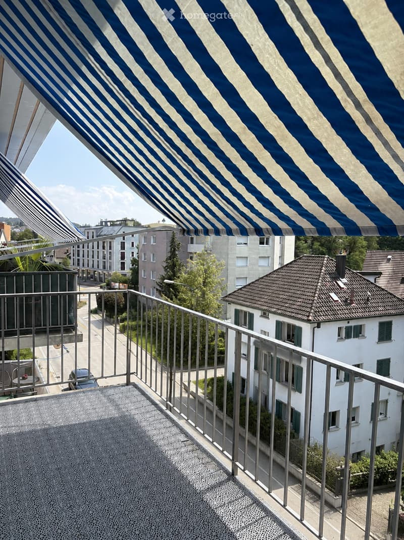 Balcony with metal railing, patterned floor, and striped awning. View of street, buildings, and greenery.