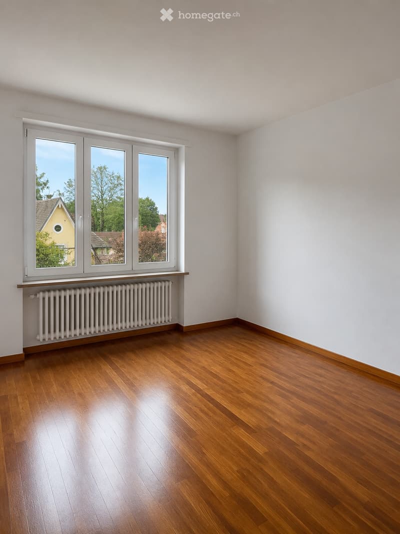 Empty room with wooden flooring, white walls, large windows, and a radiator below the windows.