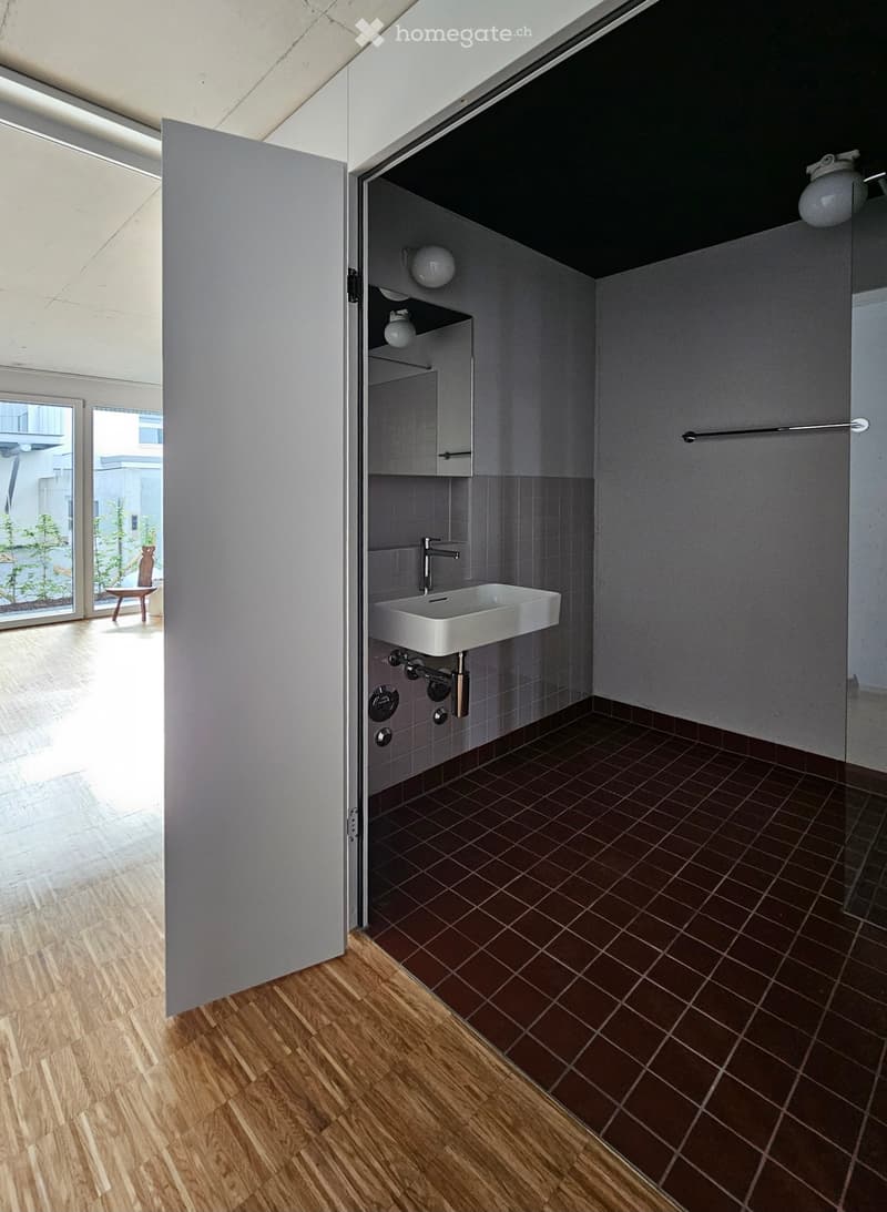 Modern bathroom with a white sink, chrome faucet, grey tiled walls, and a small cabinet with shelves holding a teapot and other items.