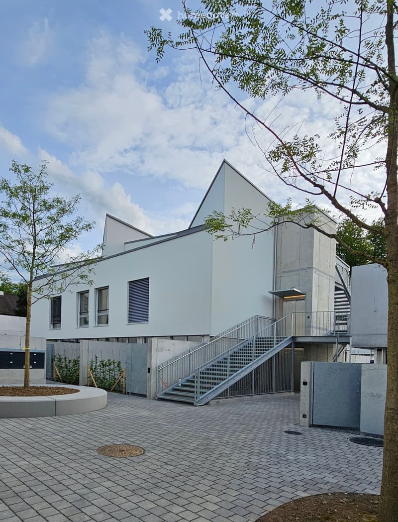 Modern two-story building with white exterior, raised ground floor, staircase leading to upper level, and surrounding paved area with trees.