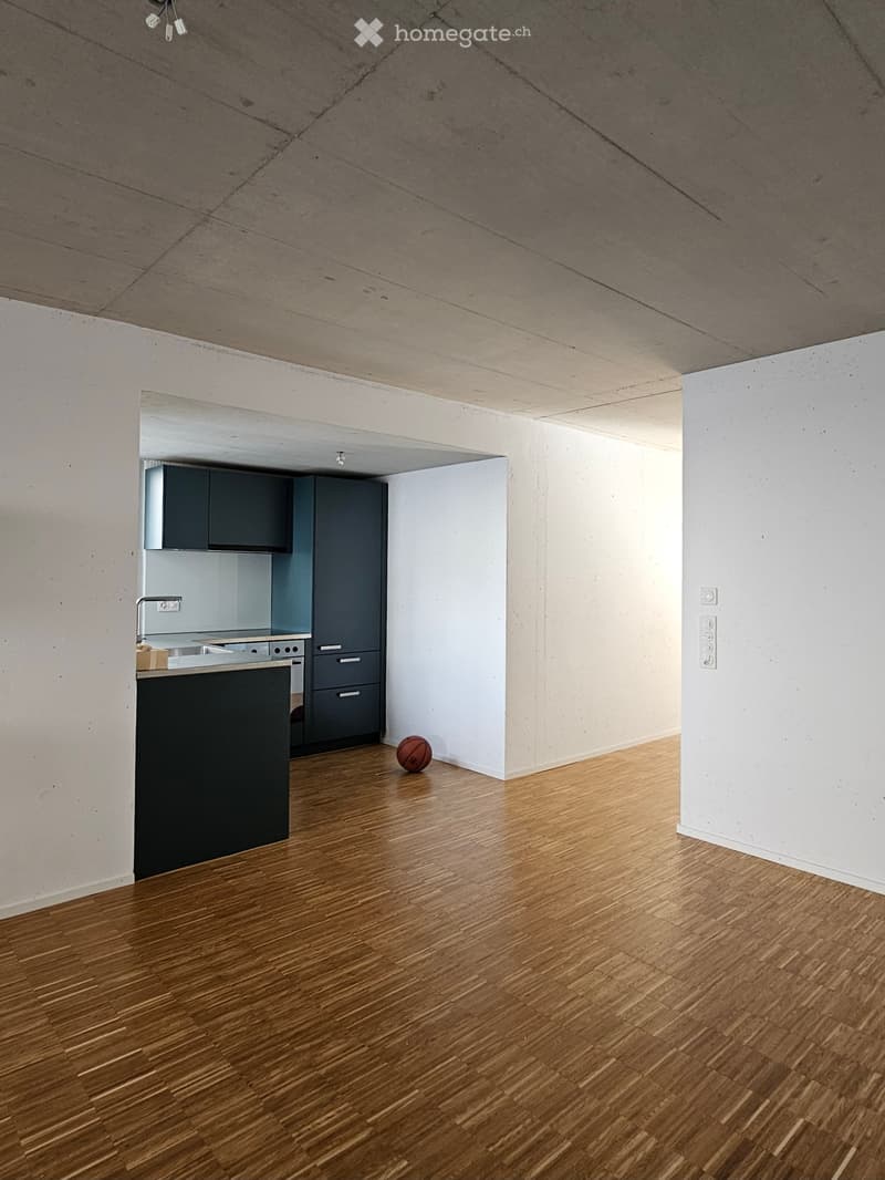Empty room with wooden flooring, white walls, and a small kitchen area with dark cabinets and stainless steel appliances.