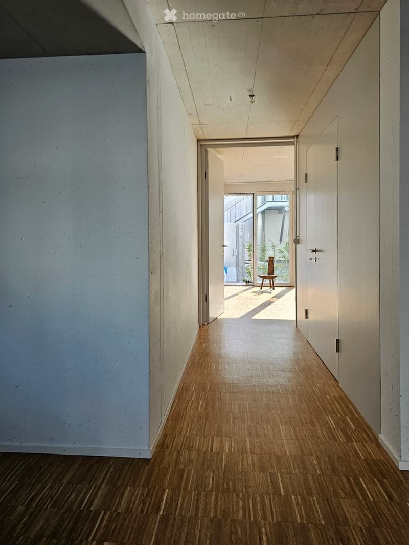 Modern hallway with wooden flooring, white walls, and recessed lighting. Features an open doorway leading to another room with visible furniture.