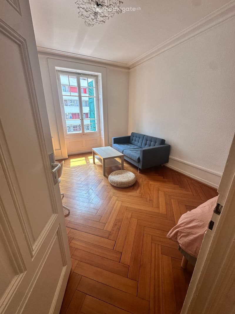 A modern living room with wooden herringbone flooring, a blue sofa, a coffee table, a round woven stool, and a bed with a pink blanket visible through an open doorway.