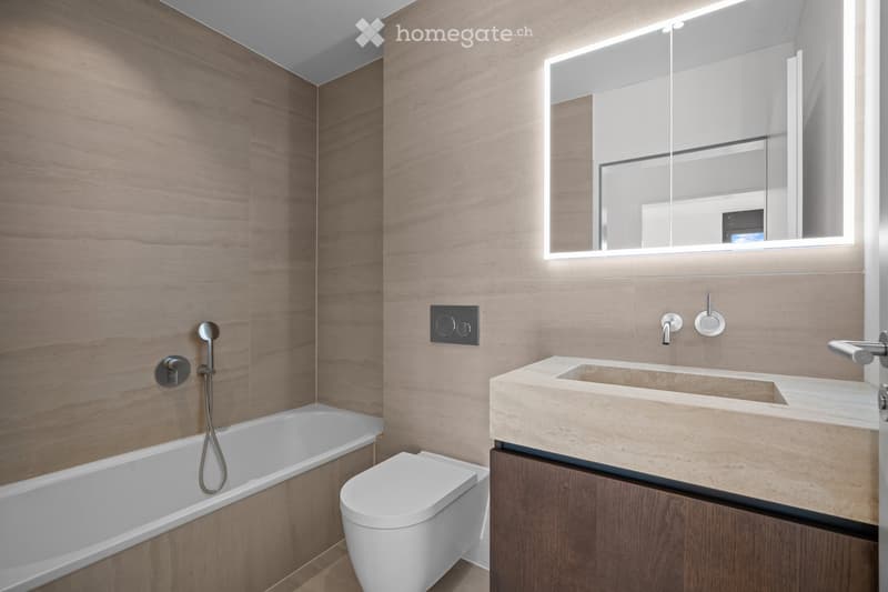 Modern bathroom with wooden walls, a bathtub, toilet, sink, and a large illuminated mirror.