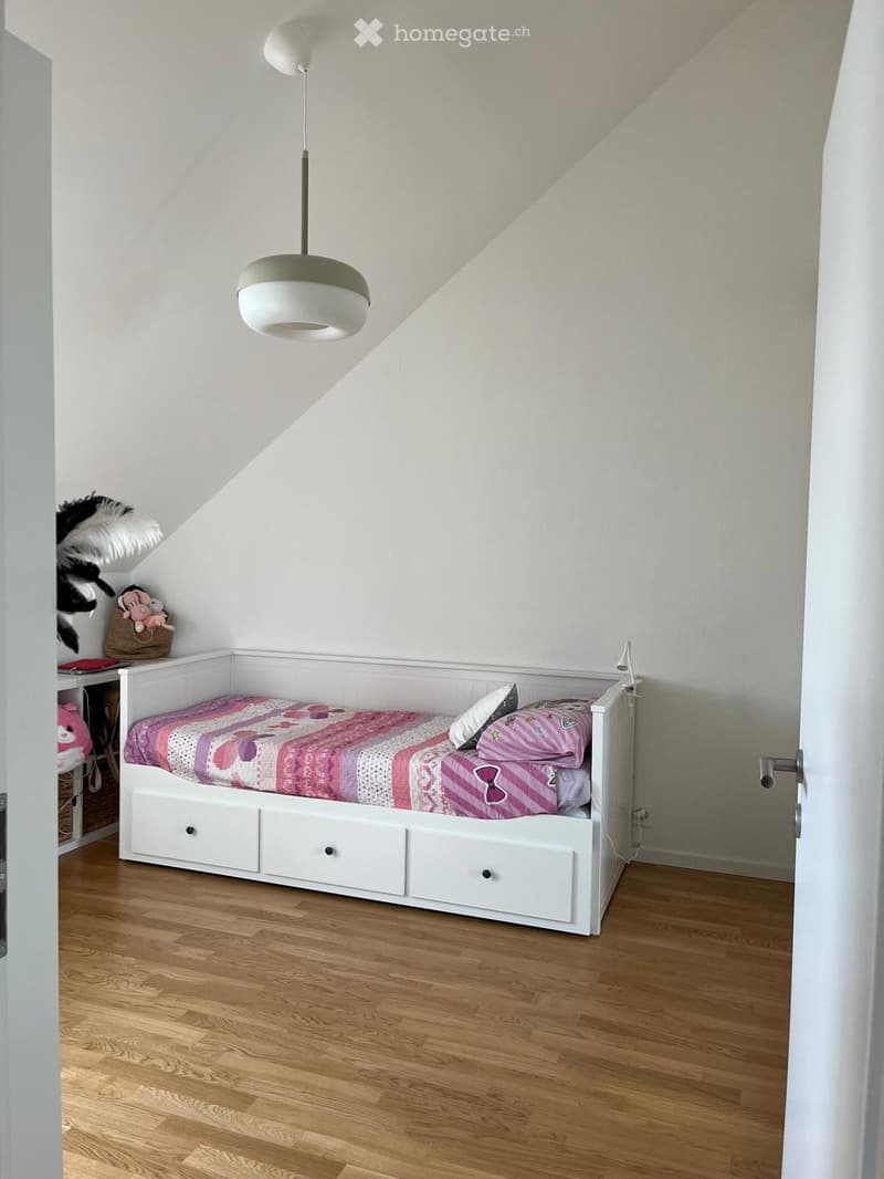 Cozy bedroom with a white bed, pink and purple bedding, a bedside table with a lamp, and a small toy on the shelf.