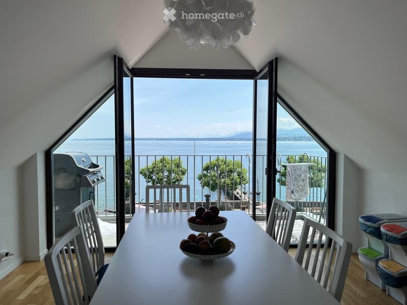 Bright room with large windows offering a scenic view, featuring a dining table with chairs and a bowl of fruit.