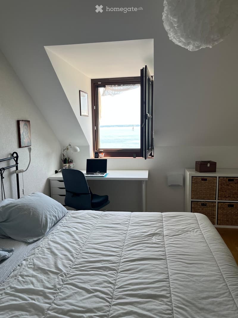 Cozy attic bedroom with a large window offering a view, a neatly made bed, a desk with a laptop, and a storage unit with woven baskets.