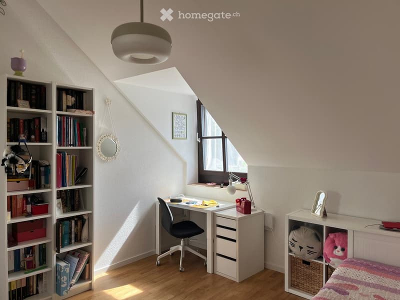 Cozy attic bedroom with wooden floors, slanted ceilings, large window, bed with colorful bedding, desk with chair, bookshelf filled with books, and decorative items.