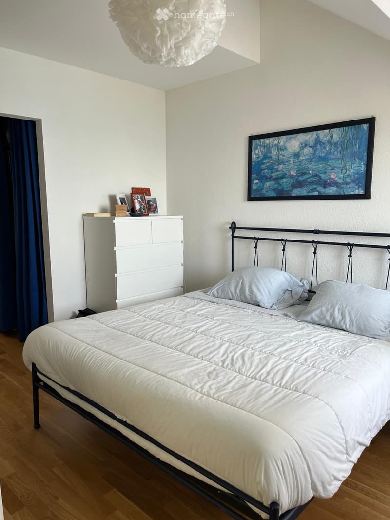 A well-lit bedroom with a neatly made bed, a nightstand, a cabinet, and a framed painting on the wall.