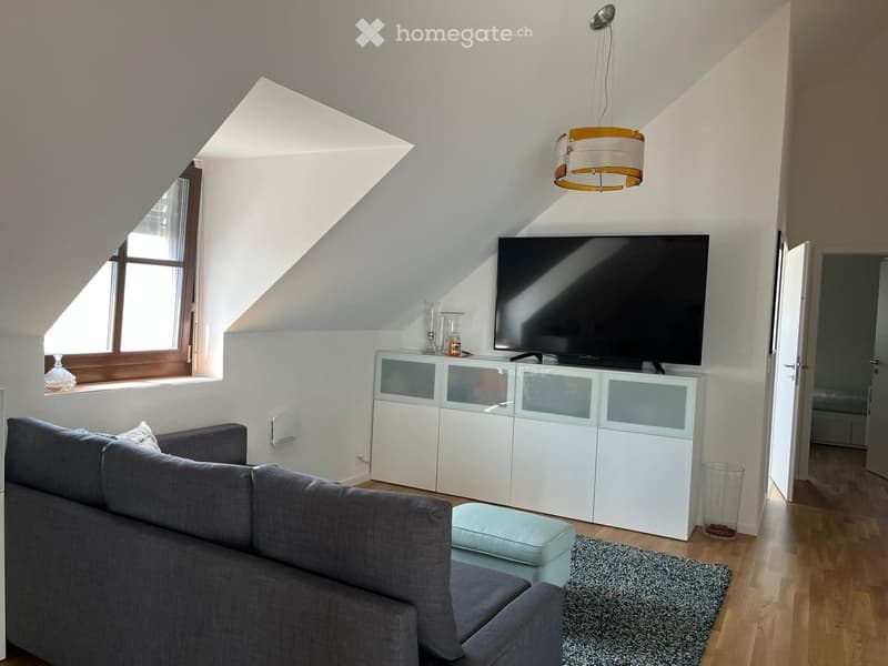 Modern living room with a grey sectional sofa, ottoman, coffee table, flat-screen TV on a white cabinet, and a hanging lamp. The room features wooden flooring, a skylight window, and a view of an adjacent bedroom.