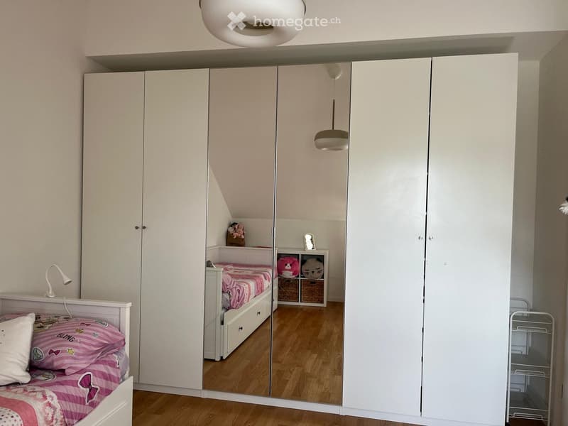 A well-lit bedroom with two single beds, white wardrobes, a large mirror, and a small nightstand with toys.