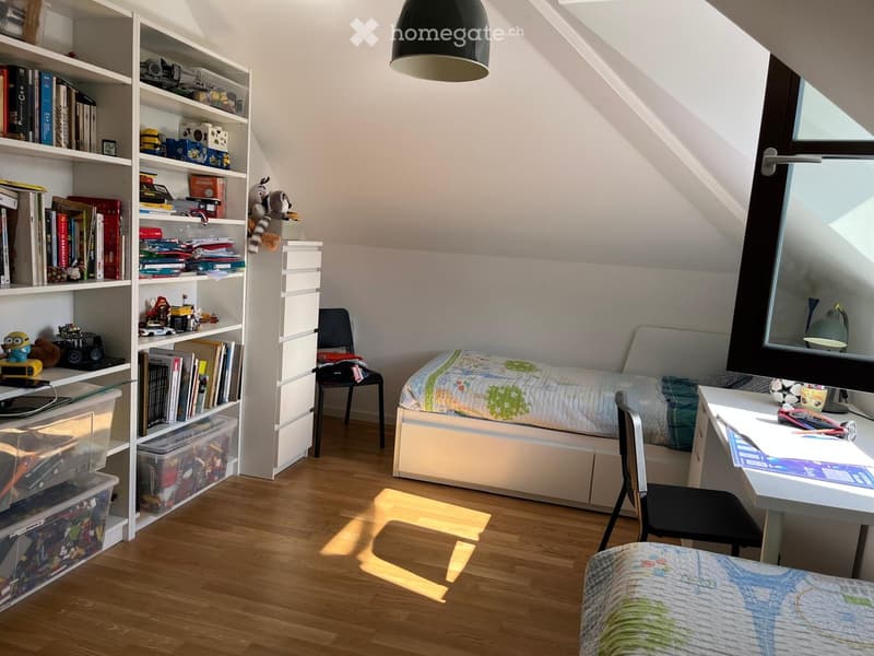 Cozy attic bedroom with slanted ceilings, wooden flooring, a single bed with colorful bedding, a desk with a chair, a bookshelf filled with books and toys, and storage bins.