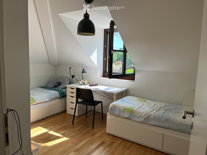 Cozy attic bedroom with two single beds, a desk, and a chair, featuring wooden flooring and large windows allowing natural light.